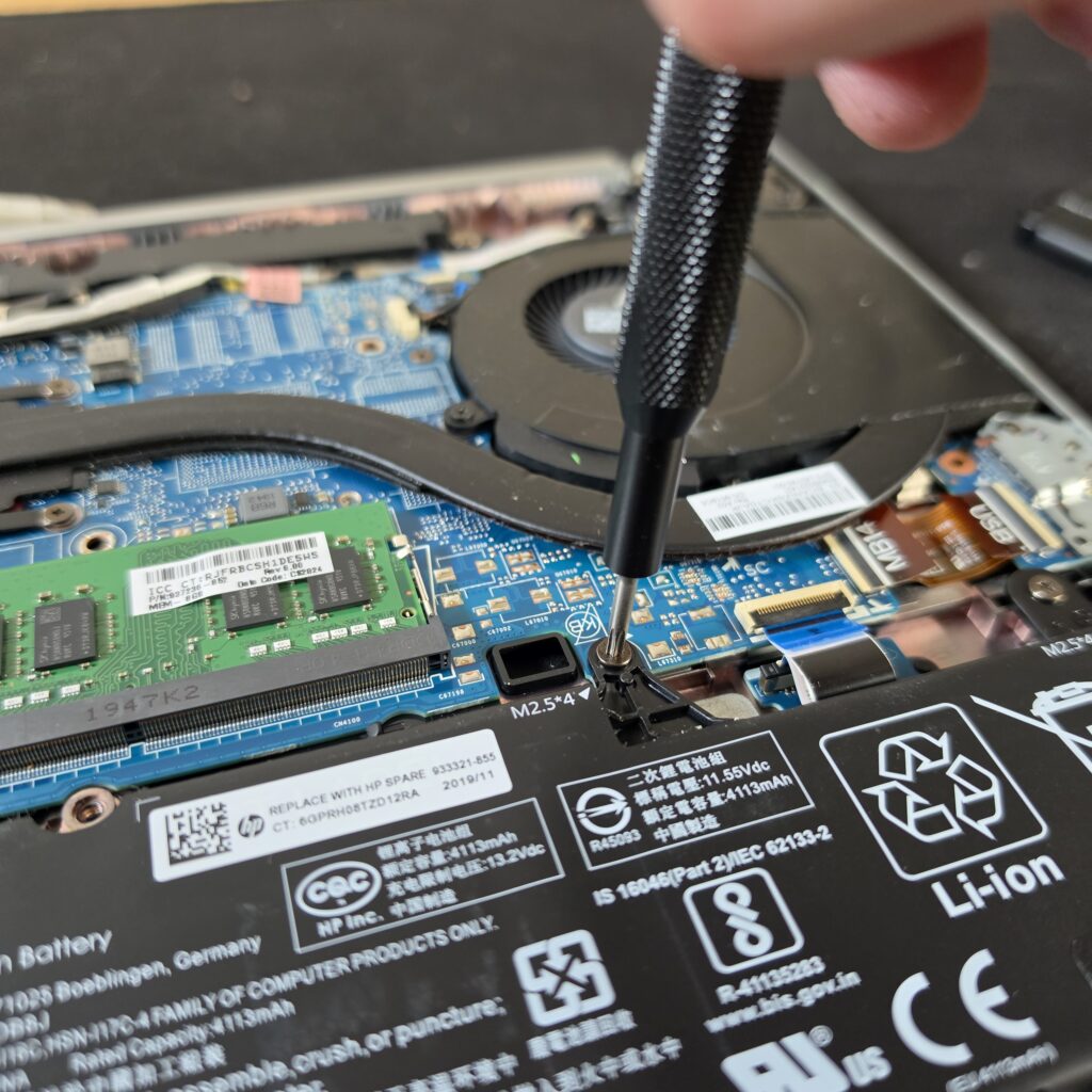Close-up of an open HP laptop showing a person using a screwdriver to remove a screw securing the internal battery, with the RAM, cooling fan, and motherboard visible.