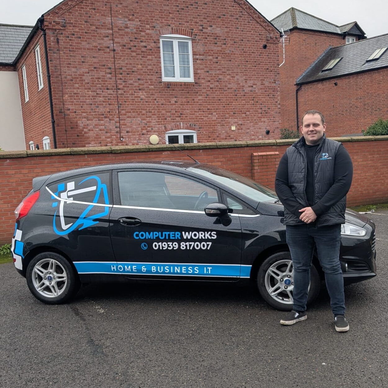 A Computer Works IT technician standing beside a black branded company car featuring the Computer Works logo, contact number 01939 817007, and Home & Business IT services signage, parked in front of red-brick buildings.